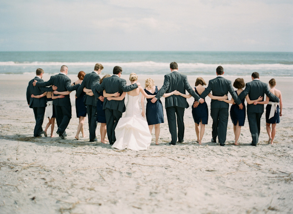 they wedding party on a beach poses with their back to the camera. Facing the water, they embrace each other- photo by South Carolina based destination wedding photographer Virgil Bunao